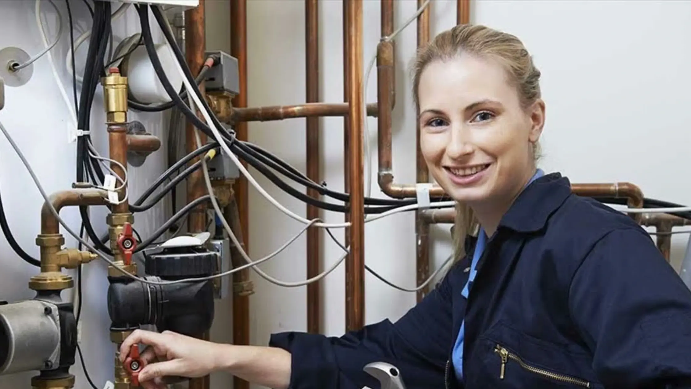 
A female plumber in dark blue overalls smiles while adjusting a valve amidst a network of pipes and wires.