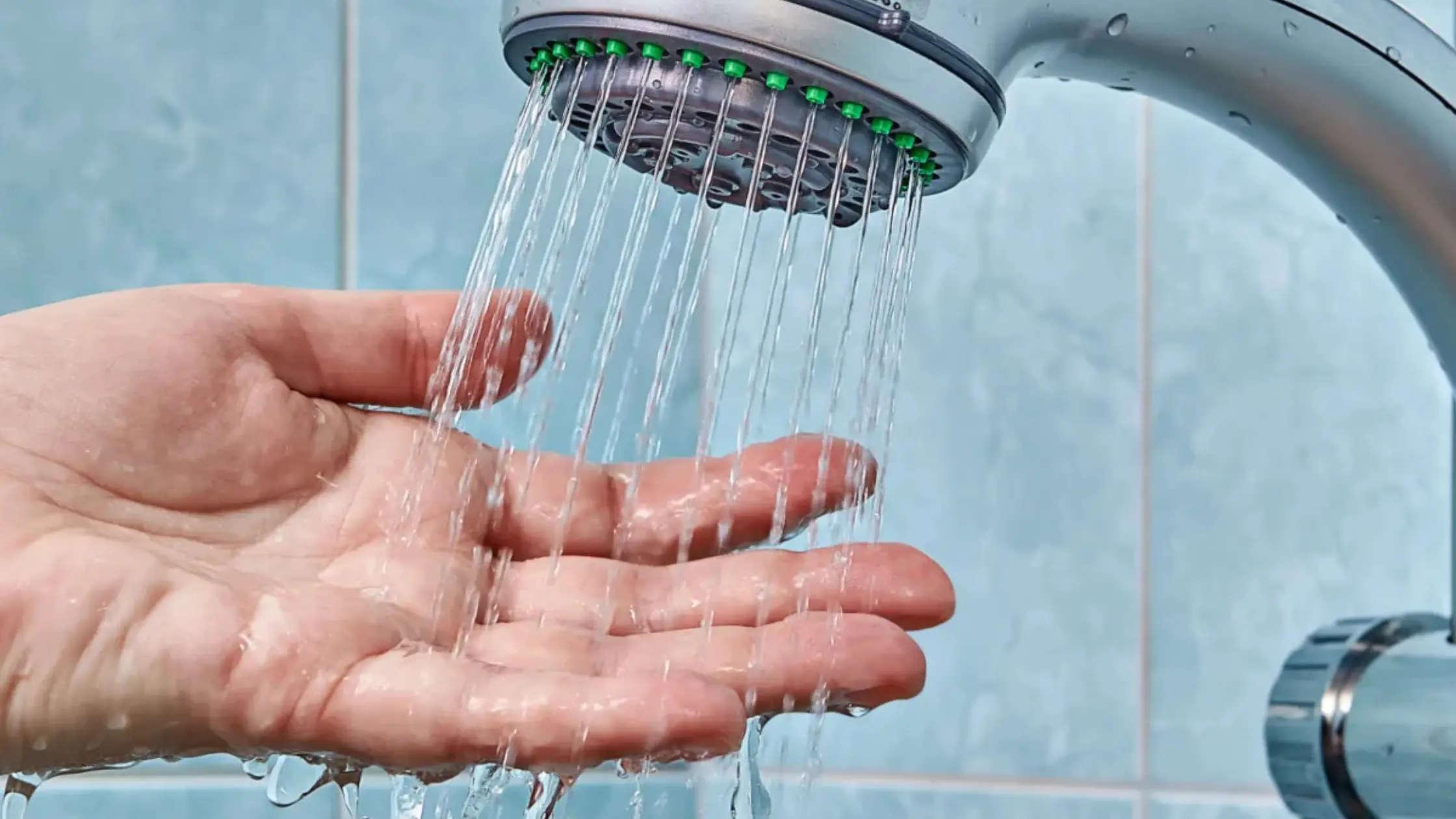  A hand is held palm up under a shower head with water spraying out, demonstrating water flow. The background is a blue tiled wall.